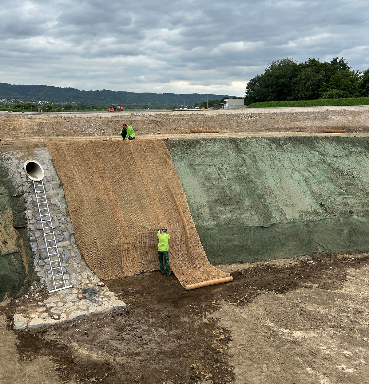 Ausführung Neisser GmbH Ein steile Böschung auf einer Baustelle, an der zwei Männer Erosionsschutzgewebe ausrollen und befestigen