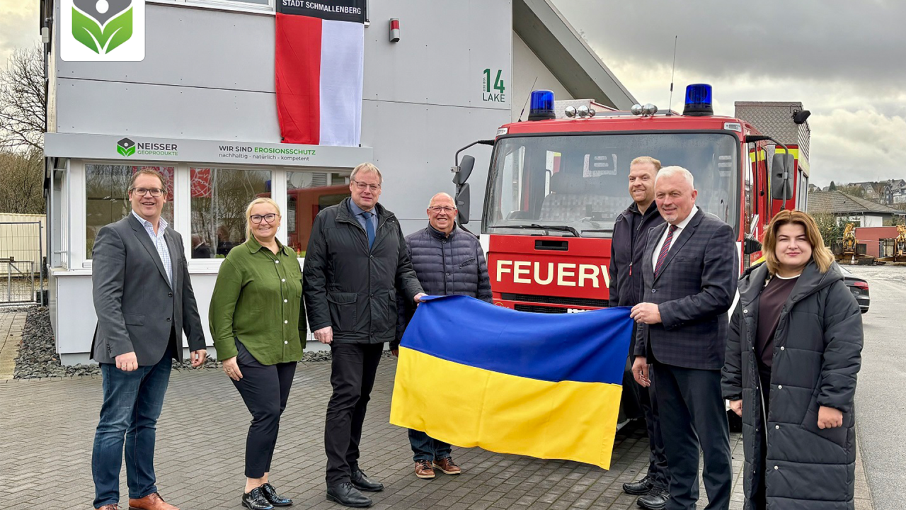 Das Ehepaar Neisser sowie der Bürgermeister der Stadt Schmallenberg, Burkhard König stehen vor dem roten Feuerwehr-Auto und halten eine Ukraine-Flagge mit Ukrainern zusammen hoch