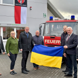 Das Ehepaar Neisser sowie der Bürgermeister der Stadt Schmallenberg, Burkhard König stehen vor dem roten Feuerwehr-Auto und halten eine Ukraine-Flagge mit Ukrainern zusammen hoch