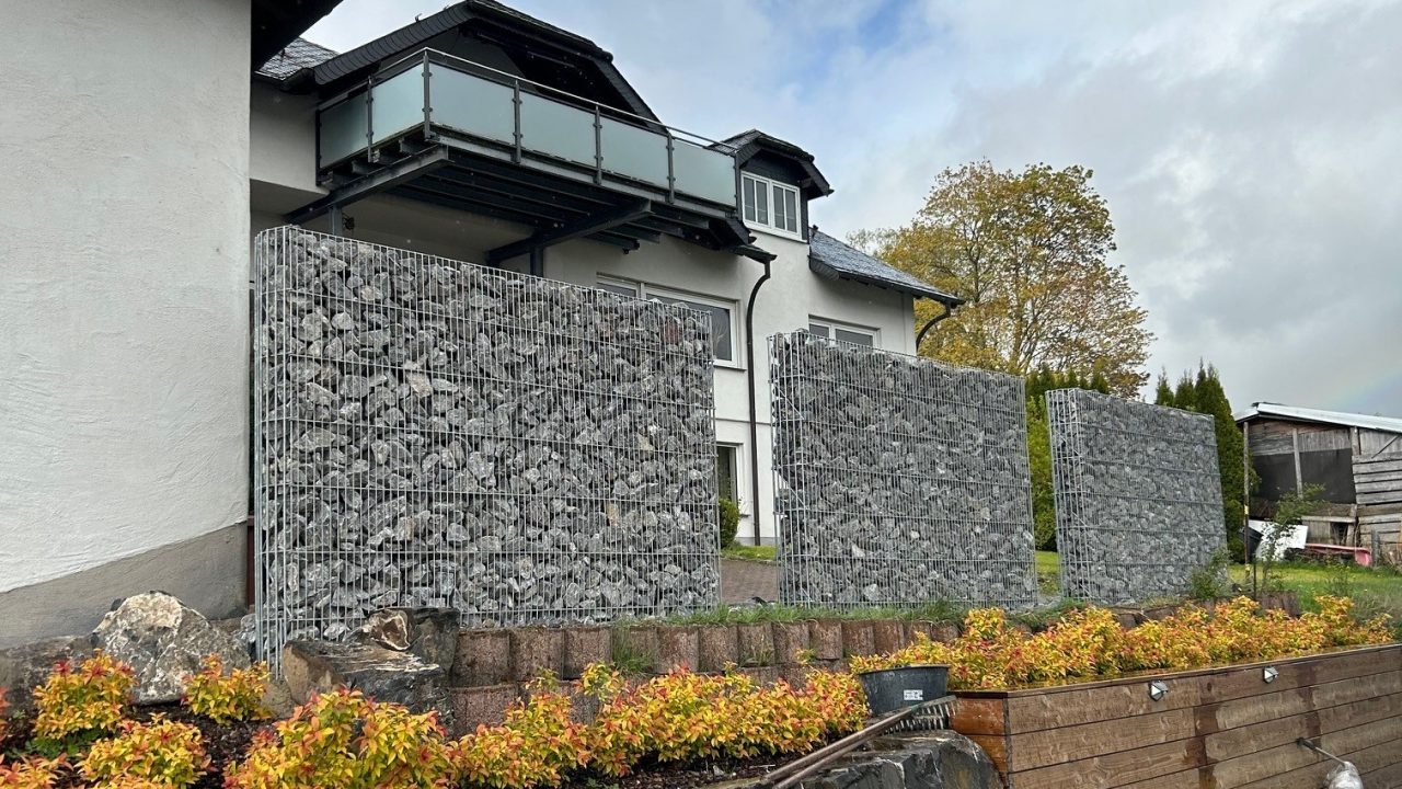 Drei Steinzaun-Elemente mit Abstand dazwischen in einem Garten, dahinter ein Wohnhaus.